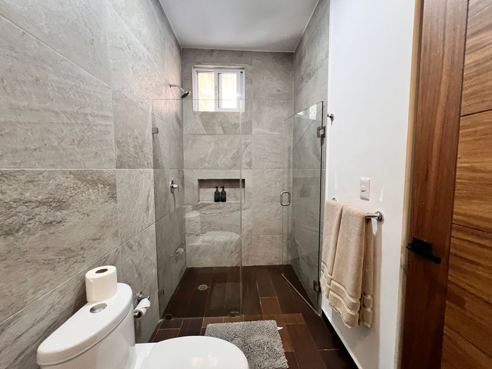 Een moderne badkamer met een glazen douchecabine, een toilet en een klein raam.