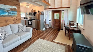 A cozy cabin interior with a living room, dining area, kitchen, and a loft bedroom with a wooden ceiling.