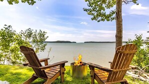 Two Adirondack chairs facing a lake with a fire pit between them.