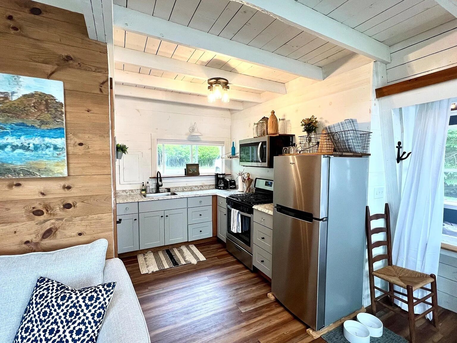 A compact kitchen with wooden cabinets, a stainless steel refrigerator, a microwave, and a stove. There's a window above the sink, a painting on the wall, and a chair by the window.