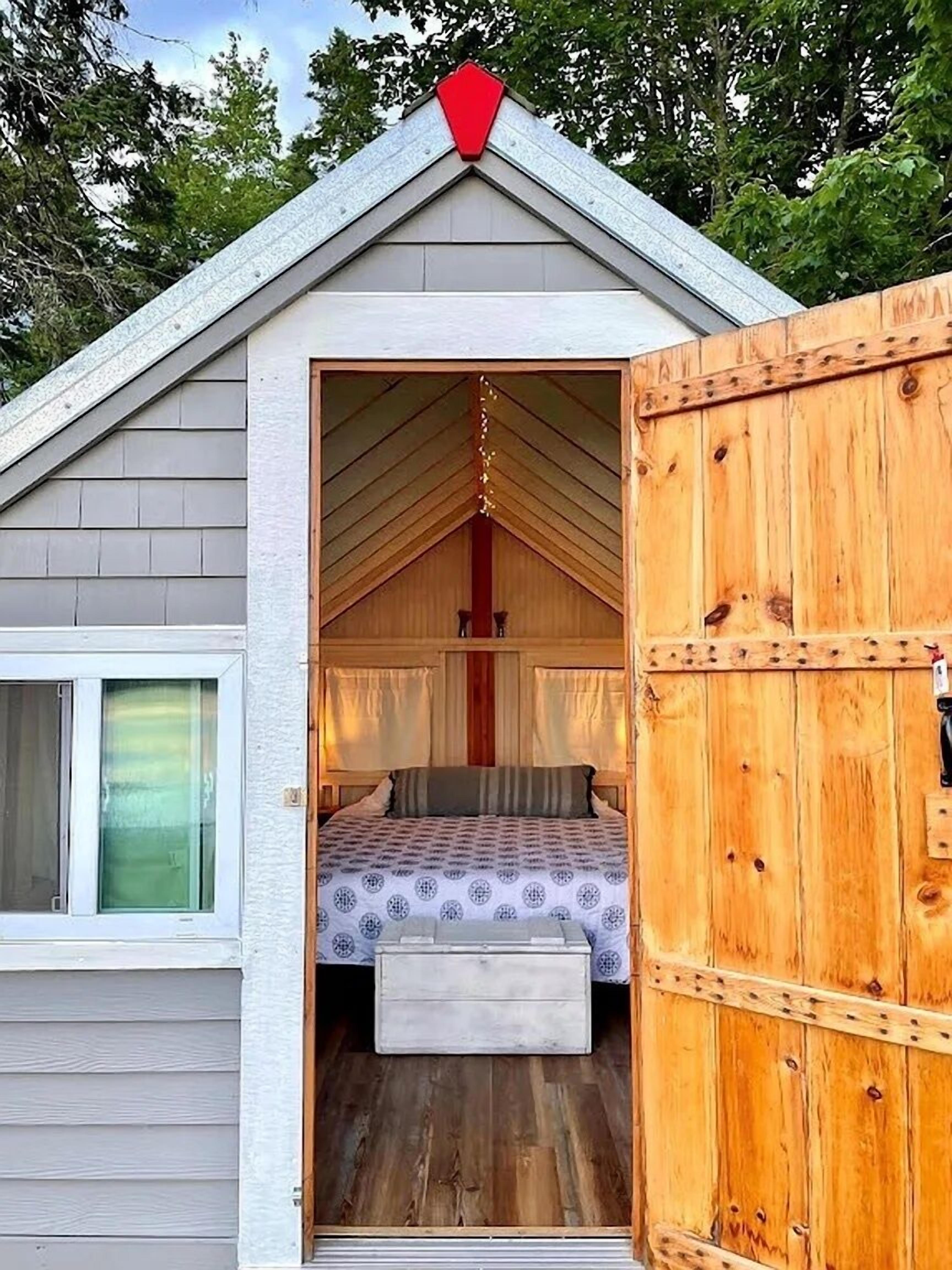 A small, cozy cabin with a sloped roof and a wooden door. Inside, there is a bed with a patterned bedspread and a small wooden bench.