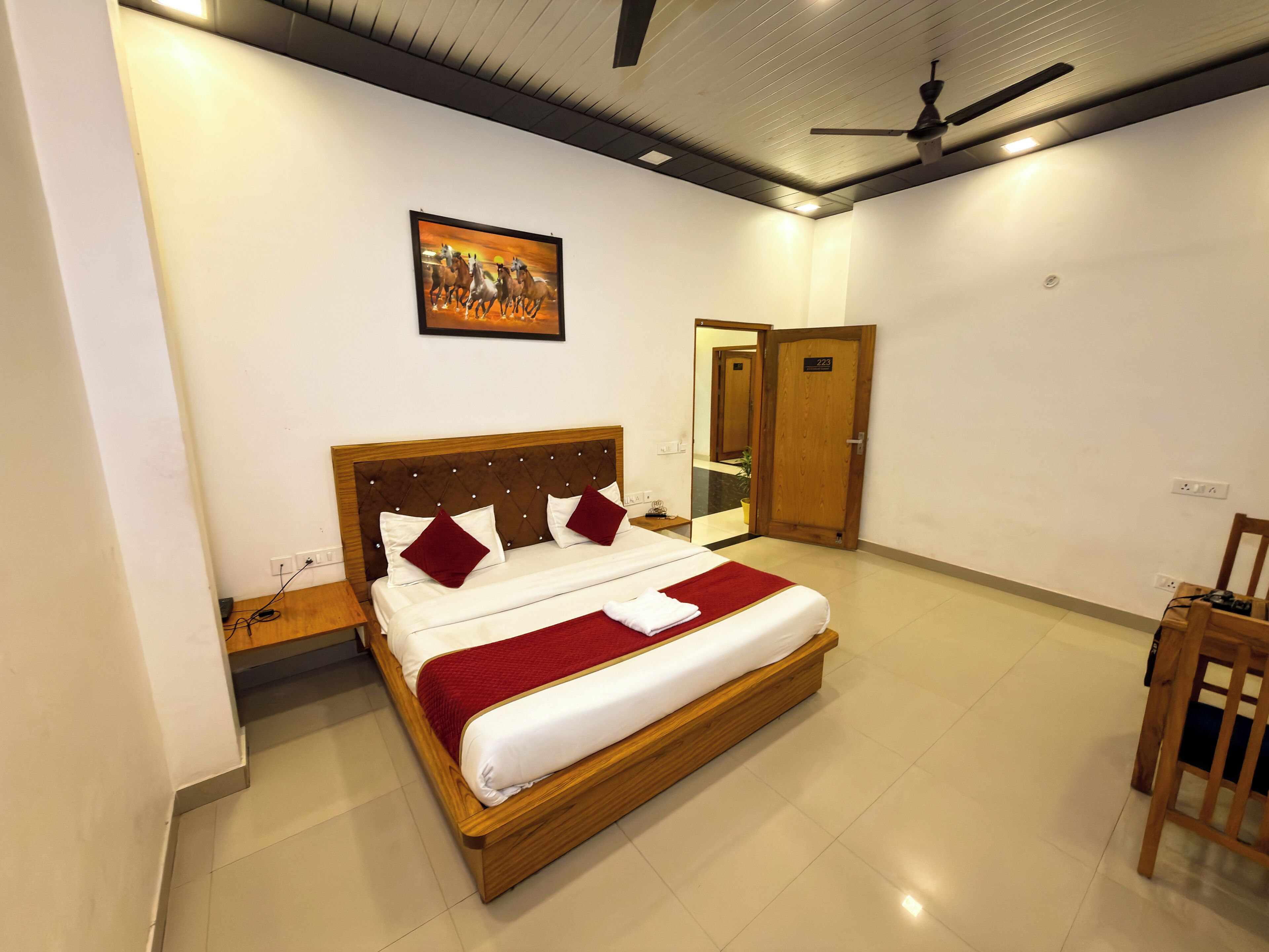 Quarto de hotel com cama de madeira, manta vermelha, ventilador de teto e um quadro na parede.