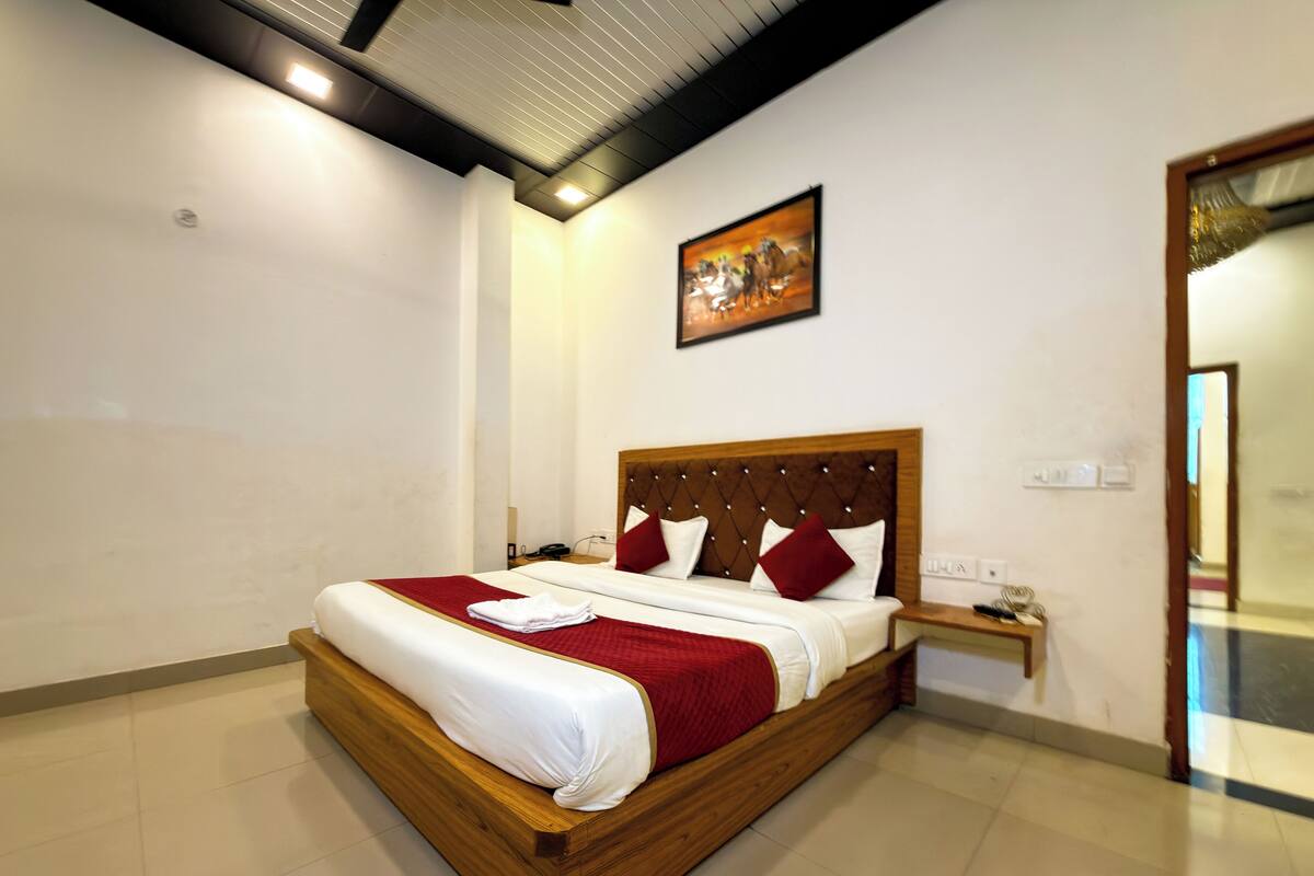 A neatly made bed with a wooden frame and red bedding, a nightstand with a phone, and a painting on the wall.