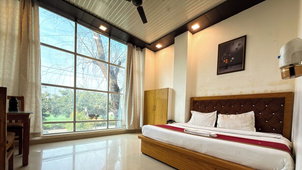 A hotel room with a large bed, a wooden headboard, a desk, and a window with a view of trees. - AARYA HOTEL & SPA Jim Corbett (Ramnagar)