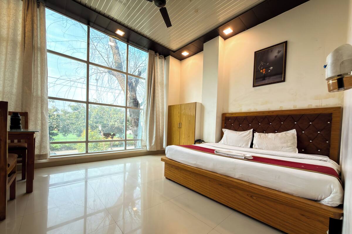 A hotel room with a large bed, a wooden headboard, a desk, and a window with a view of trees.