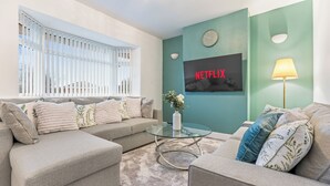 Ein Wohnzimmer mit einer Sofagarnitur, einem Glastisch, einem an der Wand montierten Fernseher mit 'NETFLIX' und einer Lampe.