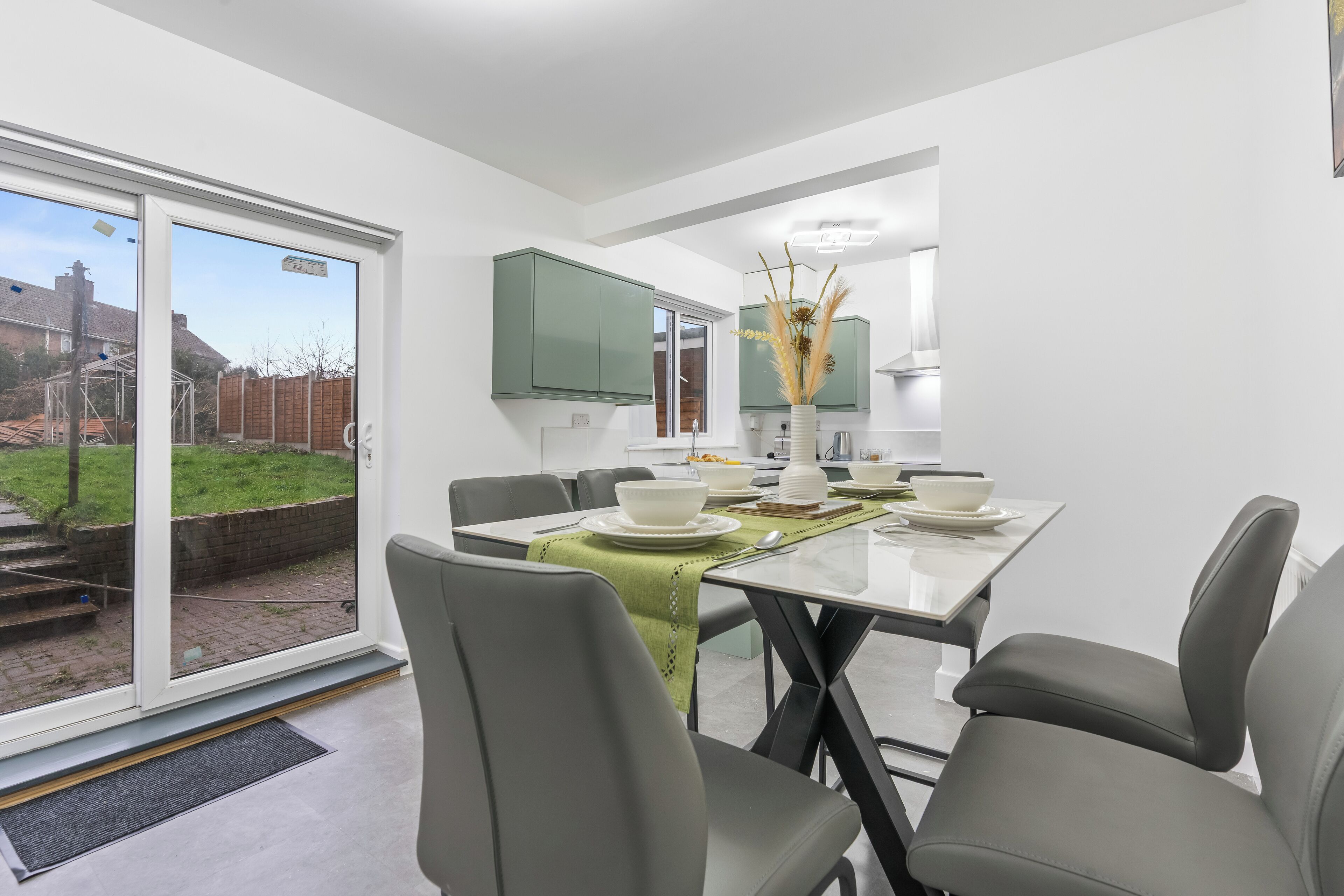 Uma sala de jantar moderna com mesa de vidro, cadeiras cinzas e vista para o quintal através de grandes portas deslizantes.