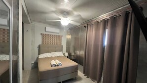 Une chambre à coucher de taille modeste, comprenant un lit, un ventilateur de plafond, une fenêtre avec des rideaux et une porte donnant sur une autre pièce.