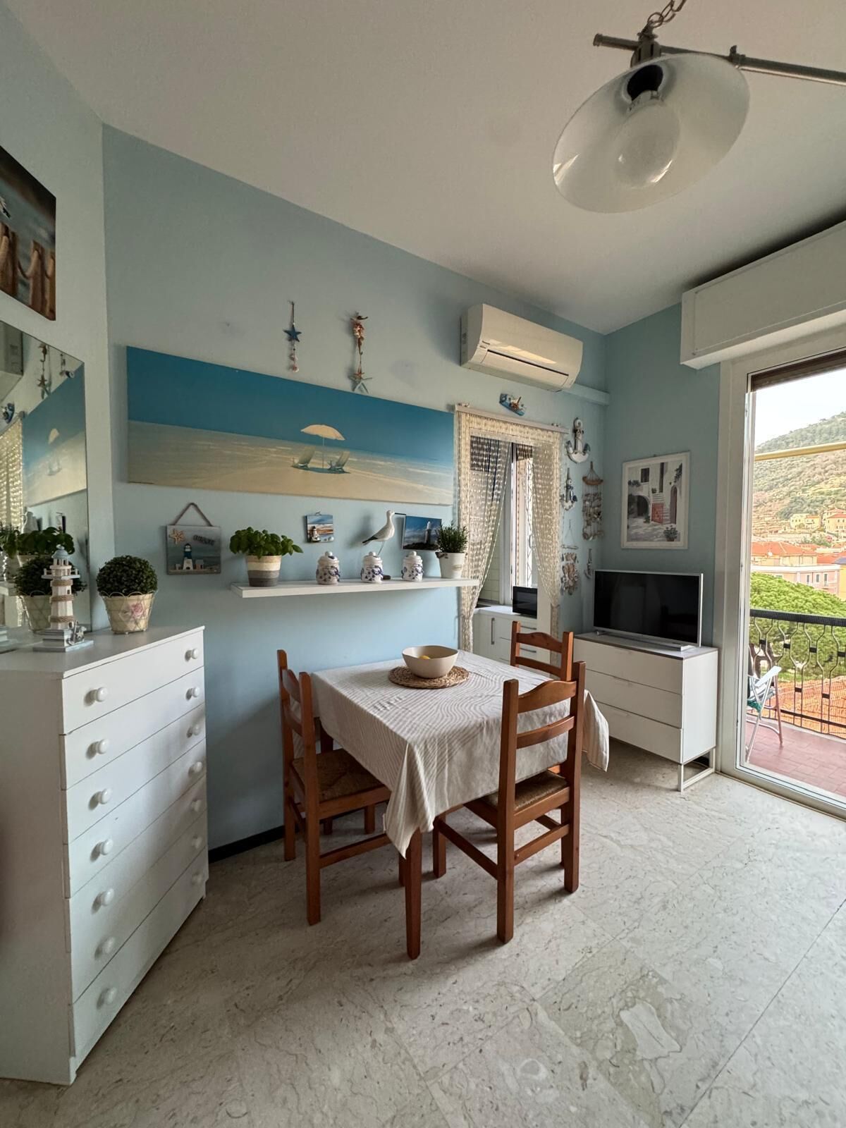A dining area with a table set for two, a white cabinet with drawers, a TV on a stand, and a view of a hillside through a balcony.