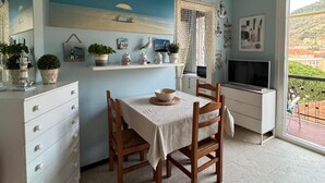 A dining area with a table set for two, a white cabinet with drawers, a TV on a stand, and a view of a hillside through a balcony. - Cozy accommodation a short walk from the sea and downtown with private parking space (Finale Ligure)