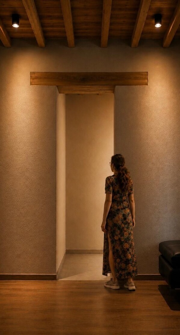 A person standing in a room with wooden beams on the ceiling, a black leather couch, and a textured wall.