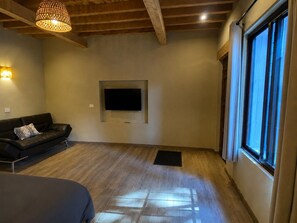 Une pièce avec un plafond en bois, un canapé en cuir noir, un téléviseur à écran plat fixé au mur et une grande fenêtre avec des rideaux.