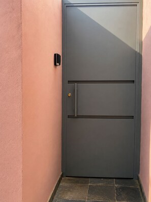 Une porte fermée, dotée d’un clavier à code et d’une poignée, se dresse sur un mur rose, devant un sol recouvert de carreaux de pierre.