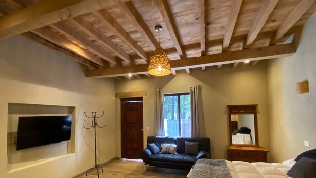 Ein Zimmer mit Holzboden, einer Holzbalkendecke, einem Flachbildfernseher an der Wand, einem schwarzen Ledersofa und einem Bett mit weißer Bettdecke.