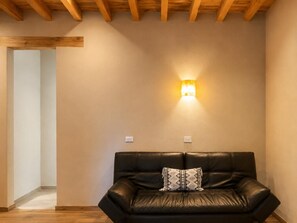 Een kamer met een zwarte leren bank, een decoratief kussen en een wandlamp, plus een houten balk in het plafond.