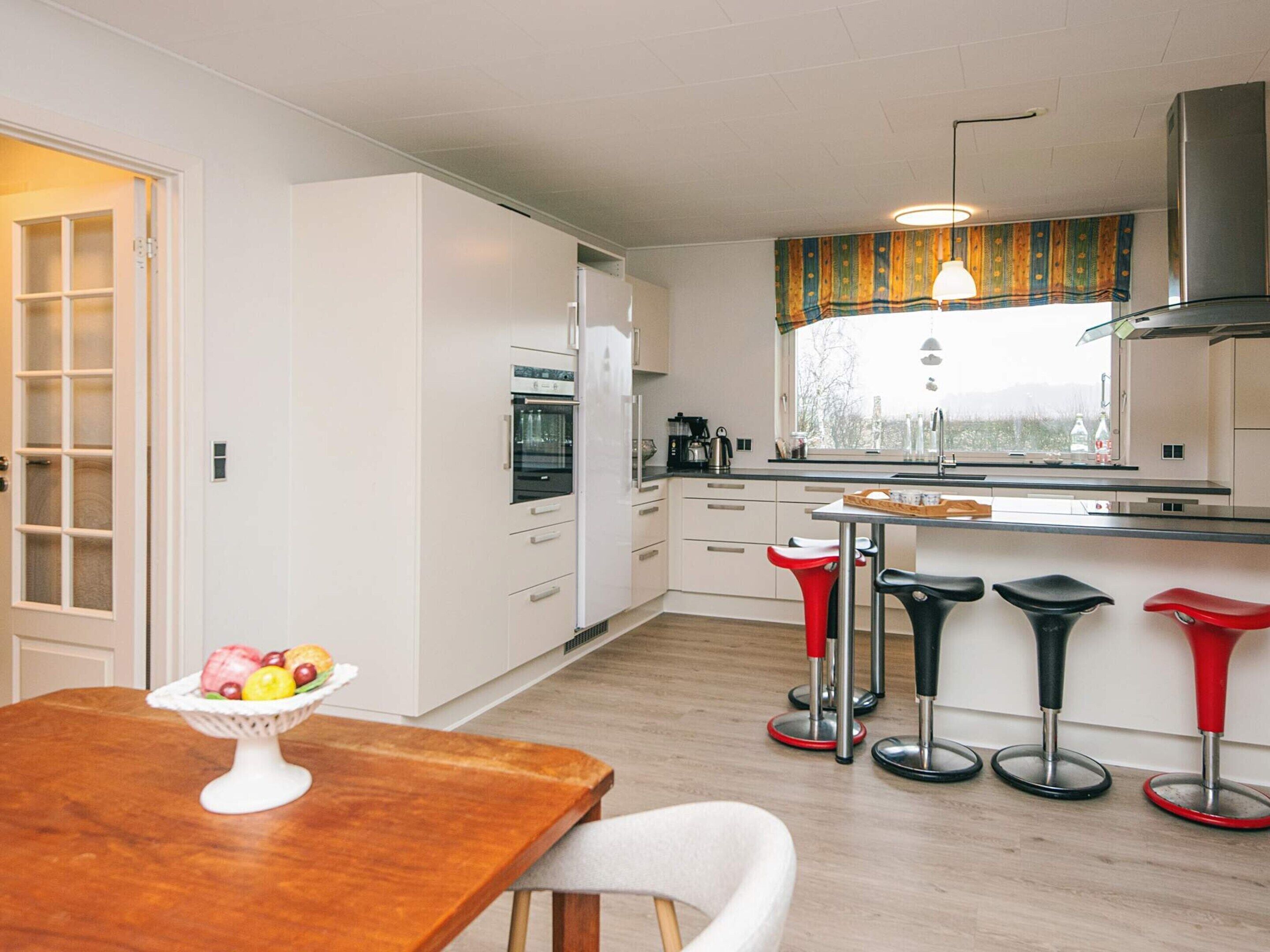 A modern kitchen with white cabinets, a built-in oven, and a window with a view of the outdoors. There is a wooden dining table with a fruit bowl and four bar stools.