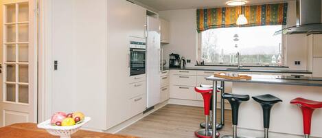 A modern kitchen with white cabinets, a built-in oven, and a window with a view of the outdoors. There is a wooden dining table with a fruit bowl and four bar stools.