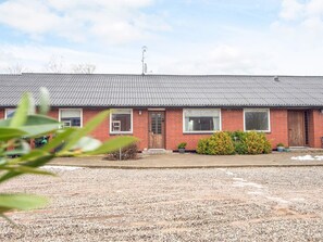 A red brick building with a metal roof, a gravel driveway, and a small garden with bushes and a potted plant. - 4 Star Holiday Home in Allingabro (Allingåbro)