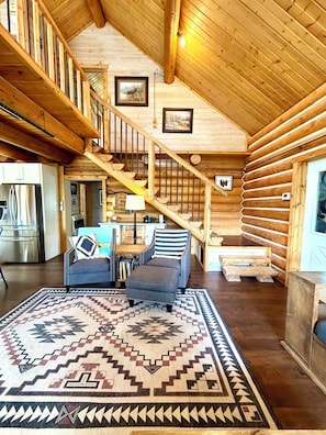 A log cabin interior with a staircase, a sofa, a rug, and a kitchen area. - Ridgeline Retreat - Fall Mountain Escape - Valley View + Fire Pit (Missoula)