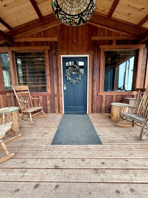 A wooden porch with a black door, a glass entry, rocking chairs, and a mosaic ceiling light.