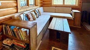 Interior de una cabaña de troncos con un banco de madera, estanterías y un ventanal con vistas al bosque.