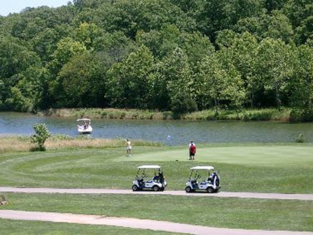 A golf course with two golf carts on a path, a lake, and dense trees in the background.