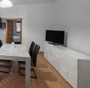 A modern kitchen with a dining area, a TV on a white cabinet, and a view into the adjacent room.