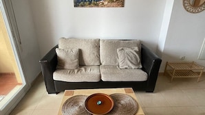 A living room with a sofa, a coffee table, and a painting of a coastal landscape. - Modern, Stylish Beach Apartment with Stunning sea views, Pools & Ultrafast Wi-Fi (La'Azohia)