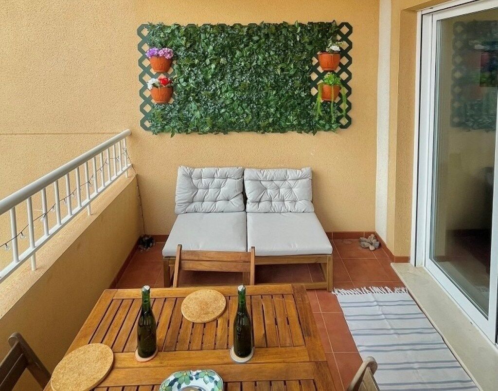 Un balcón con una mesa de madera, dos sofás acolchados y un muro decorativo de hojas verdes.