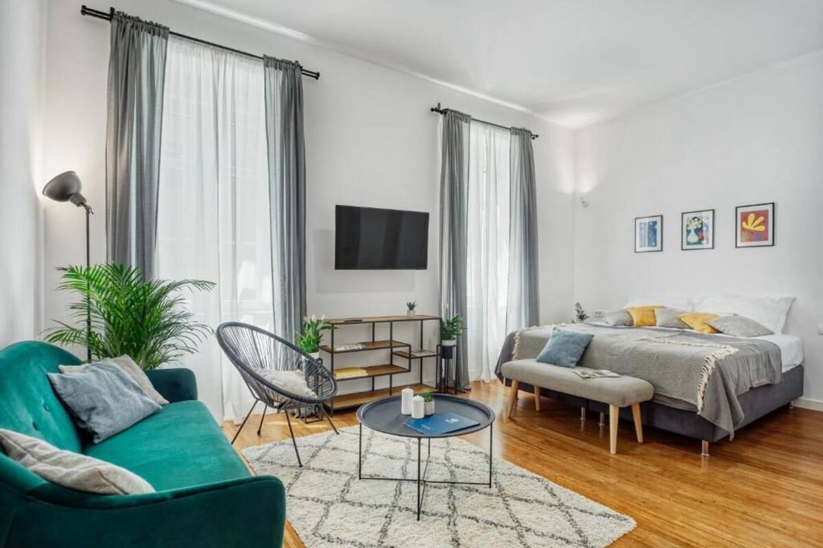 Ein modernes Wohnzimmer mit einem grünen Sofa, einem runden Couchtisch, einem schwarzen Sessel, einem Bett mit grauer Bettwäsche und einem an der Wand montierten Fernseher.