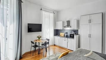 A compact room with a bed, a small dining table with chairs, a kitchenette, and a TV mounted on the wall.