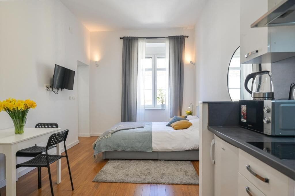 A compact bedroom with a bed, a small desk with a chair, a television on the wall, and a window with curtains.