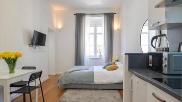 A compact bedroom with a bed, a small desk with a chair, a television on the wall, and a window with curtains.