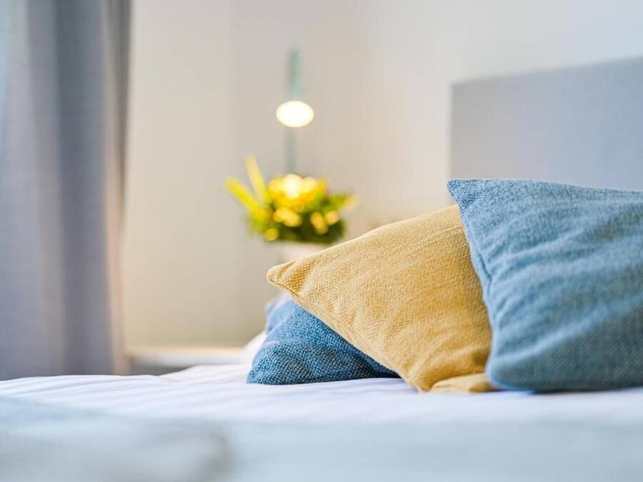 A bed with blue and yellow pillows, a nightstand with a lamp, and a flower arrangement in the background.