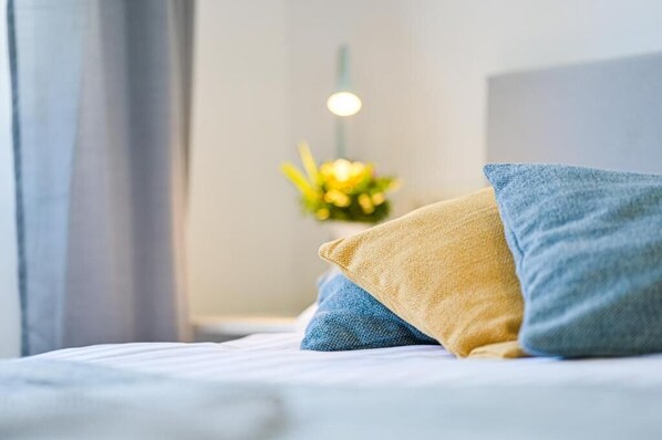 Una cama con almohadas azules y amarillas, una mesita de noche con una lámpara y un arreglo floral al fondo.