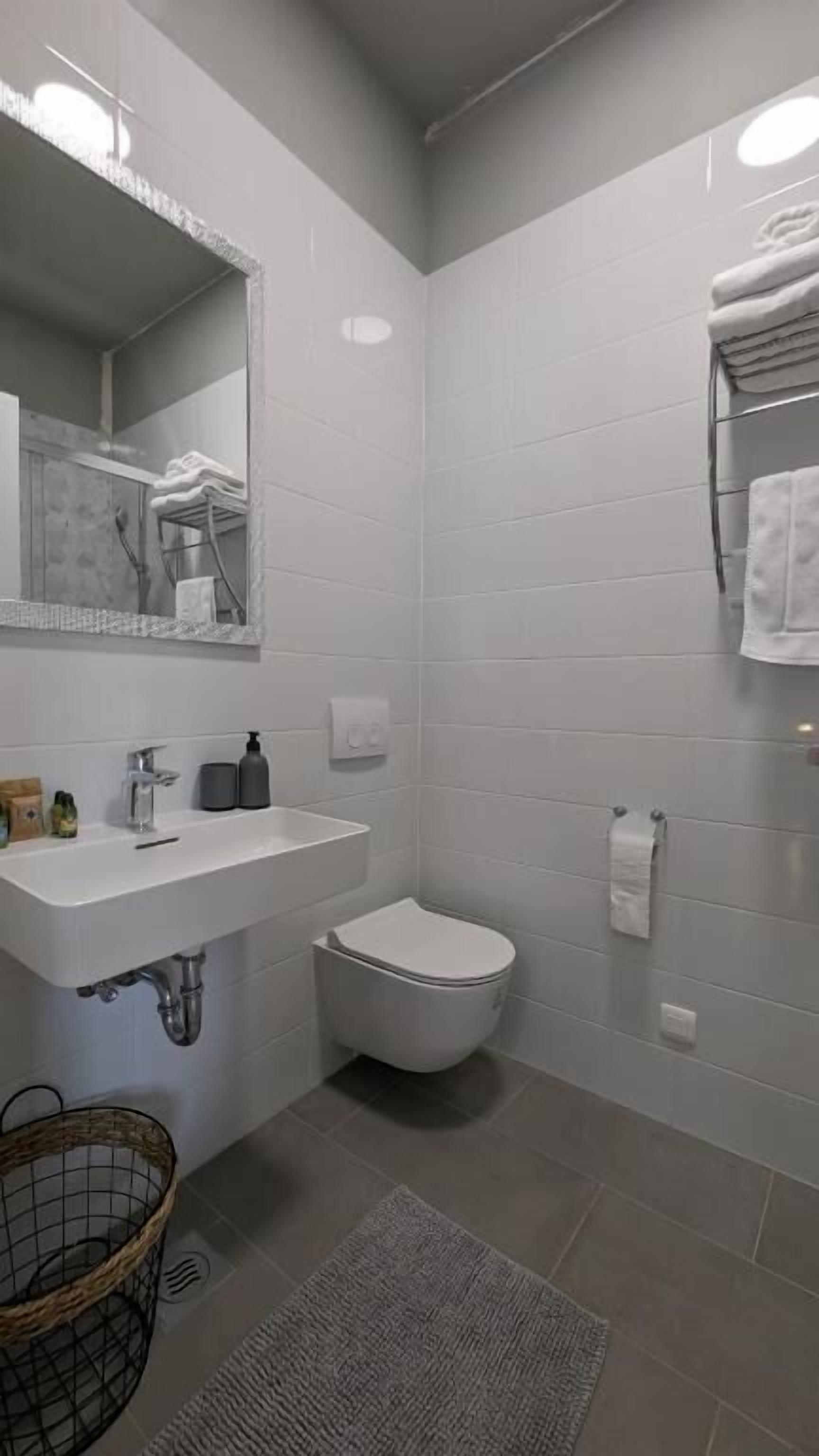 Une salle de bain avec un lavabo blanc, un miroir et des toilettes. Il y a un porte-serviettes au mur et un petit panier sur le sol.