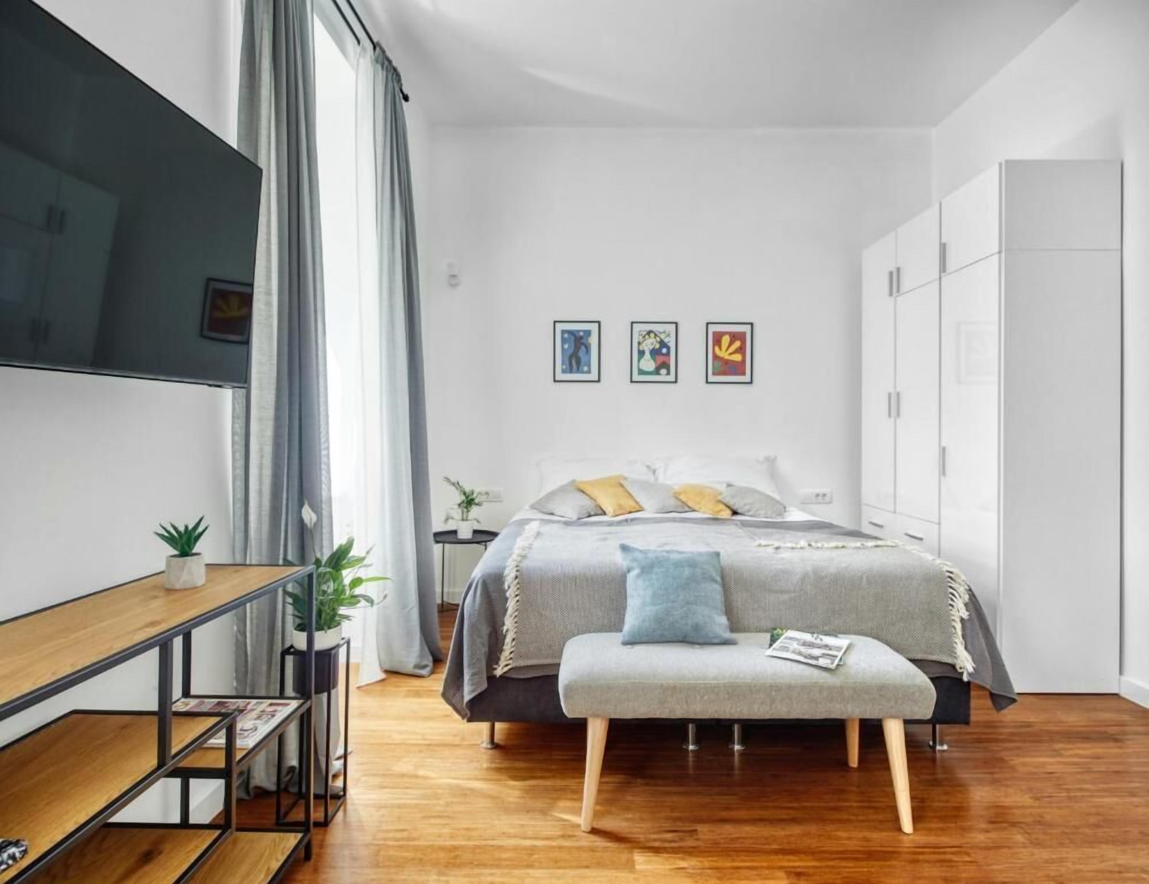 Un dormitorio moderno con cama, mesita de noche y un banco. Hay tres cuadros enmarcados en la pared, una planta en maceta y un armario.