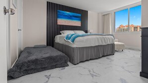 Una habitación de hotel moderna con una cama grande, un sofá pequeño, vistas de la ciudad y un cuadro en la pared.