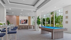 Un área de descanso moderna con mesa de billar, asientos, un televisor y ventanas grandes con vistas a palmeras.