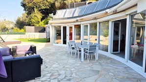 Ein überdachter Patio-Bereich mit Steinboden, Gartenmöbeln und einer Glasveranda mit Schiebetüren.