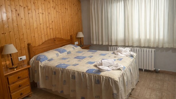 A bedroom with a wooden headboard, a bed with a blue and white patterned bedspread, two bedside lamps, a radiator, and a window with sheer curtains. - Turisme Rural El Janpere - La Cabanya (Sant Joan de les Abadesses)