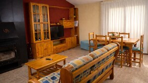 A cozy living room with a fireplace, wooden entertainment center, sofa, and dining table. - Turisme Rural El Janpere - La Cabanya (Sant Joan de les Abadesses)