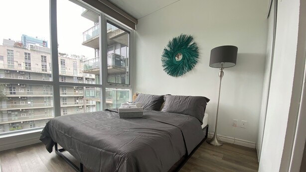 Ein Schlafzimmer mit einem großen Fenster, einem Bett mit grauer Bettwäsche, einer Stehlampe und einem dekorativen Kranz an der Wand.