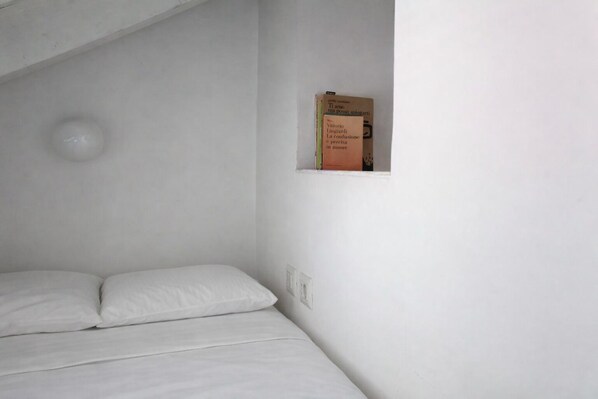 A minimalist room with a single bed, a small shelf with books, and a light fixture. - Corsico by Wynwood House (Milan)