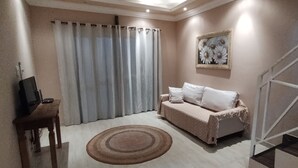 A living room with a sofa, a TV on a wooden table, a large window with curtains, and a painting on the wall. - Complete house with a beautiful view of nature. (Bananal)