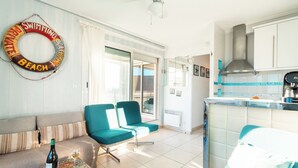A living room with a sofa, two teal chairs, a small table with a bottle and glass, and a kitchen area with cabinets and a sink. - Terrasses Catalanes Sea&Mountain View near the beach & private parking (Canet-en-Roussillon)
