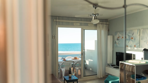 A room with a view of the sea, a dining area with chairs and a table, a sofa, a small table with a bottle and glass, and a bathroom with a sink and mirror. - Terrasses Catalanes Sea&Mountain View near the beach & private parking (Canet-en-Roussillon)