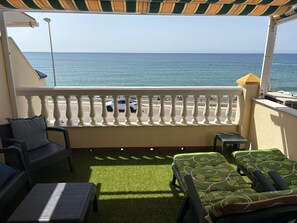Un balcon avec vue sur la mer, une voiture blanche garée dans la rue, et du mobilier d’extérieur comprenant une chaise et un canapé.