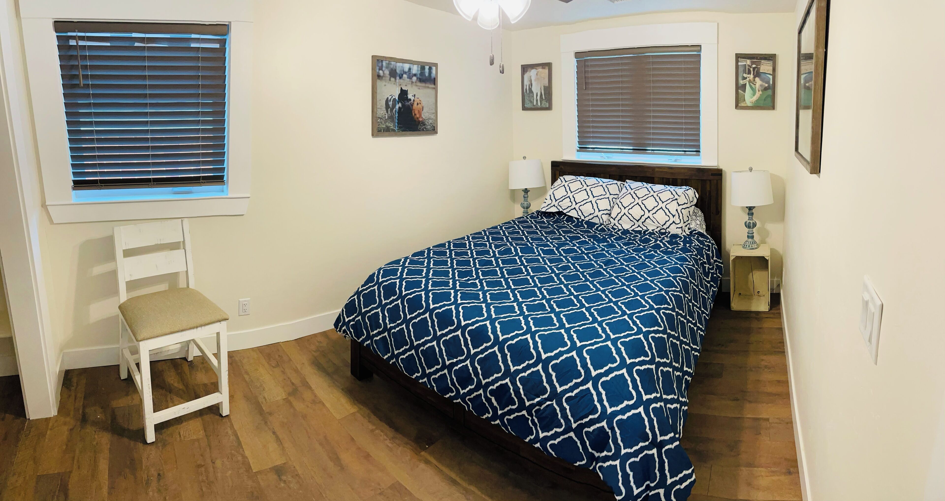 A bedroom with a bed, two bedside lamps, a chair, and two windows with blinds.