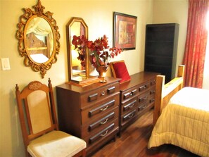A bedroom with a wooden dresser, two chairs, a bed, a mirror, and a framed picture on the wall.